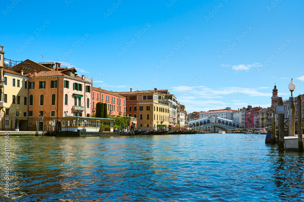 Beautiful cityscape in Venice, Italy. Picturesque old buildings surround the iconic Rialto Bridge on the Grand Canal, accompanied by gondolas and boats under vivid blue sky on sunny spring day.