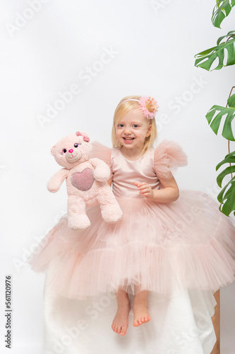 Cheerful girl in elegant dress and pretty hoop holds teddy bear. White background and houseplant leaves. Vertical frame.
