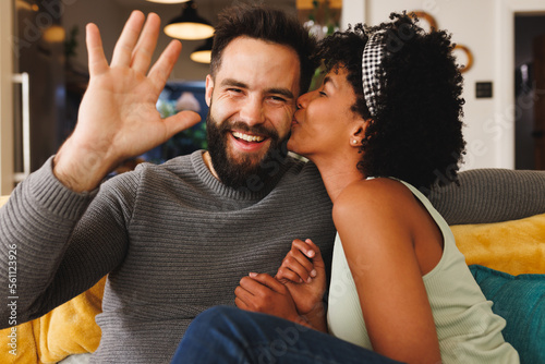 Happy bearded biracial man waving hand and girlfriend kissing him on cheek while relaxing on sofa