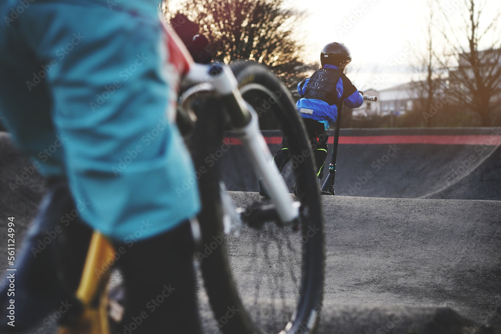 small boy with protective gear has fun with a stunt scooter on an modern asphalted pump track in bright evening light. bicycle following him. selective focus. modern sport activity concept