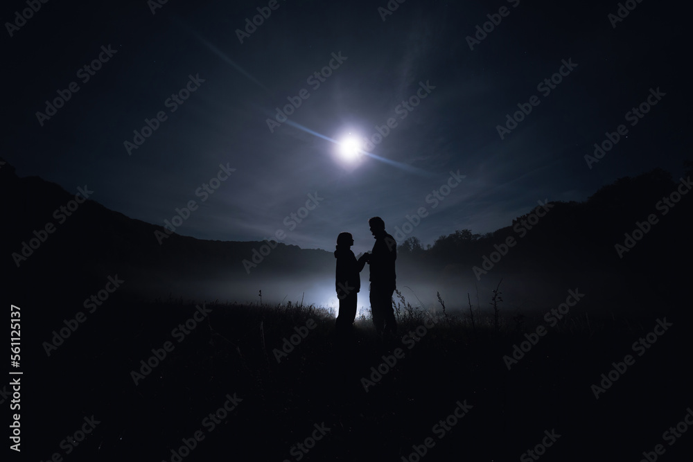 Silhouette of lovers. A couple of people, a man and a woman, are standing in the middle of a thick fog that is creeping over the ground. Mist is mystical in the moonlight. Field night