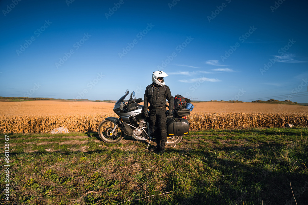 Obraz premium A man, a motorcycle driver, stands next to his touring motorcycle. Large side bags for luggage. A field of dry yellowed corn. Freedom and travel transportation concept.
