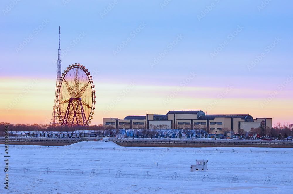 Ferris wheel and TV tower on the bank of a snow-covered river on a winter morning. Sunrise at the International Border between China and Russia
