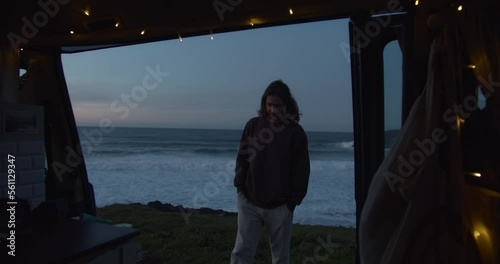 Young man watching the sunset and waves seen out of  a van with fairy lights around the door in Galicia, Spain