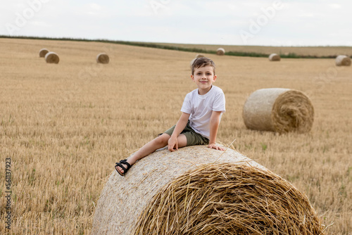 A cute little boy in a white t-shirt and khaki shorts is sitining on round bales of hay. Photo session in the field