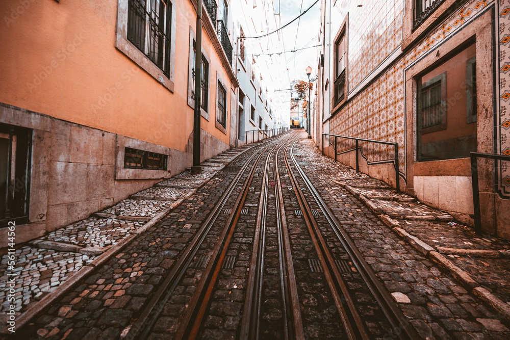 A wide-angle view of a road with a retro two-lane tramway over paving stone on a narrow street in a picturesque old neighborhood in shades of salmon on the walls and tiles in Lisbon, Portugal