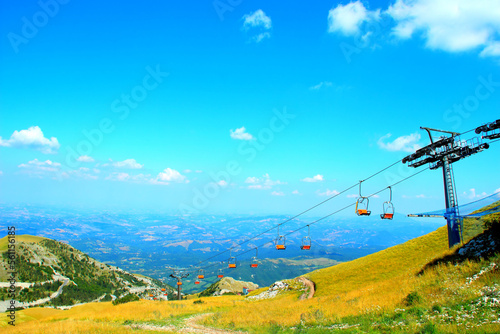 Sublime view from Sassotetto at the wondrous Marche landscapes in the slightly hazy background and a sturdy chairlift with orange seats in the foreground above green grass on a fine summer day photo