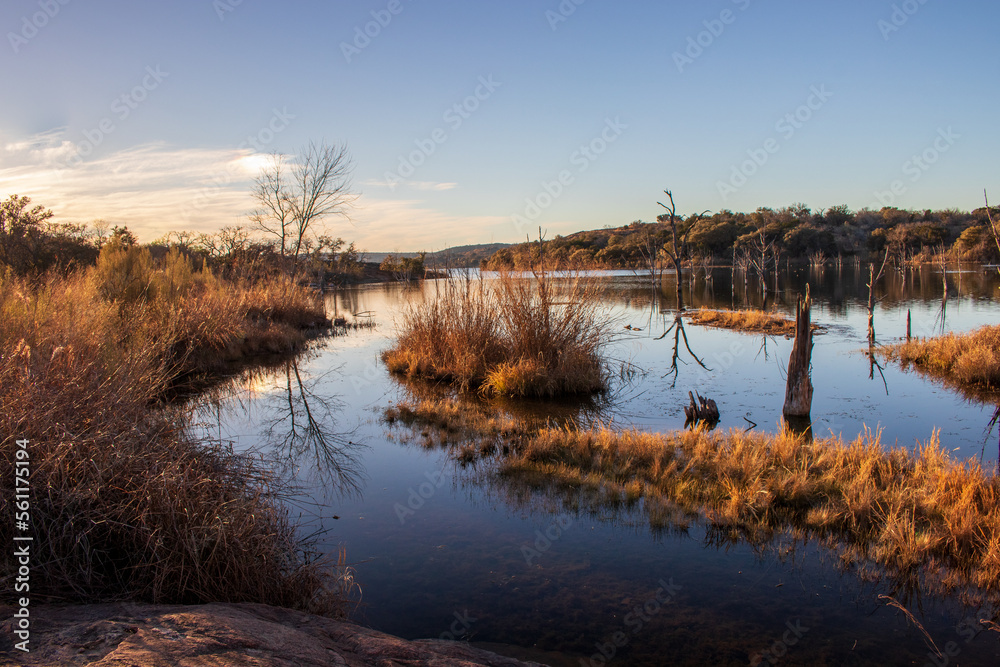 Fototapeta premium Golden hour sunset over a lakeshore changing the colors of the trees and grass to yellow and orange. Many people visit Inks Lake State Park in Texas during the Fall and early Spring