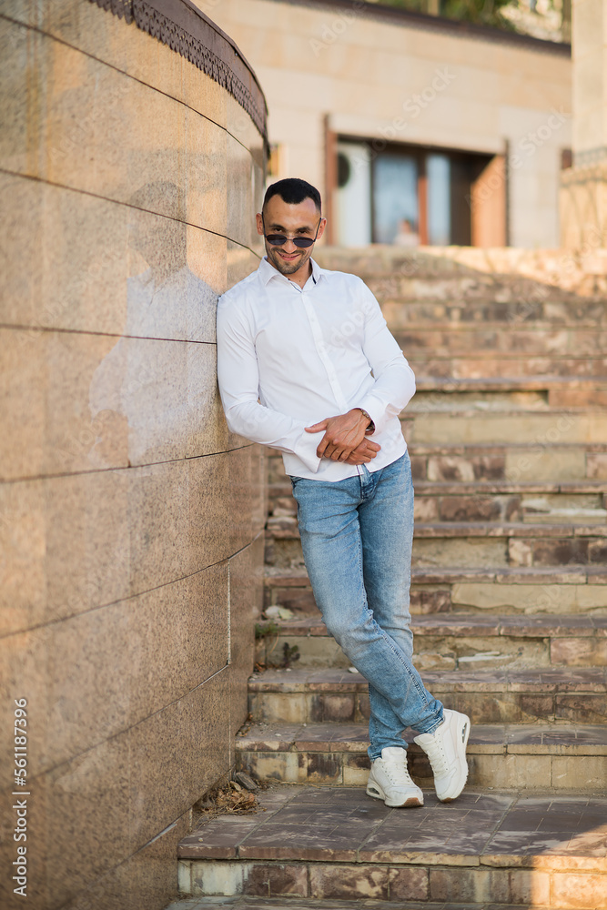 Obraz premium Handsome man in a trendy white shirt and sunglasses in the city. Handsome and charming man posing outdoors, urban style