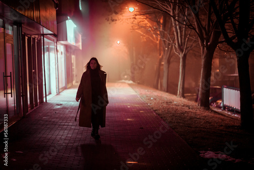 a lonely young woman in a coat walks through the night foggy city of fields lit by lampsм