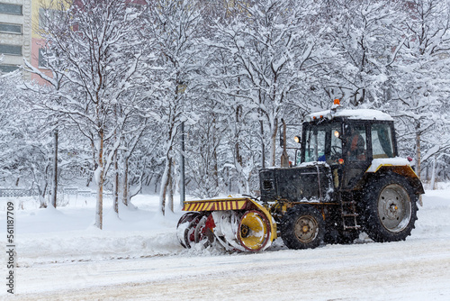 Snow plow tractor clears snow from the street during a snowfall. Snow-covered city street. Cold snowy winter weather.