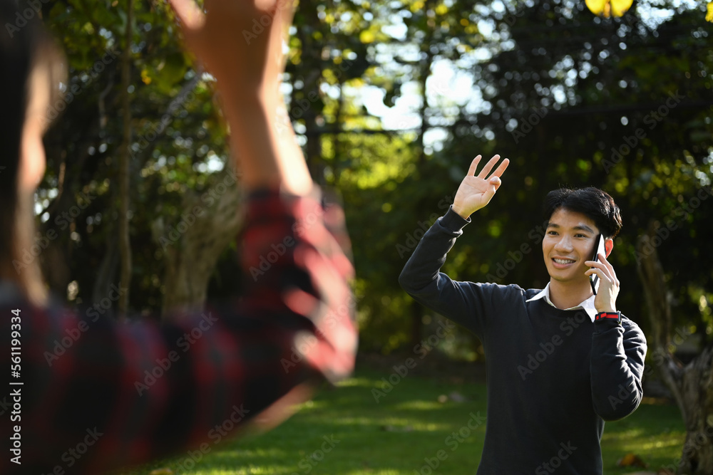 Friendly asian man walking in public park with mobile phone and greeting friend with hand raised up