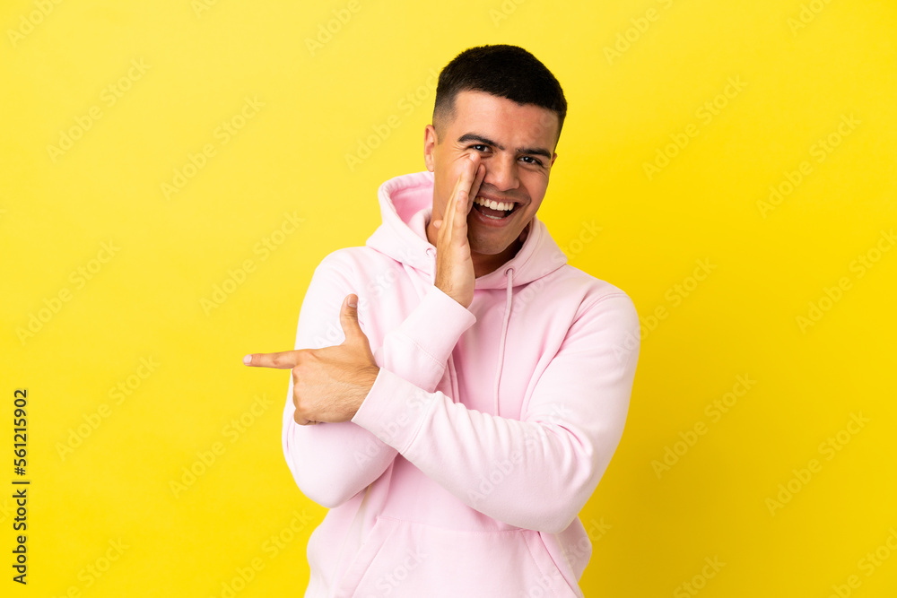 Young handsome man over isolated yellow background pointing to the side to present a product and whispering something