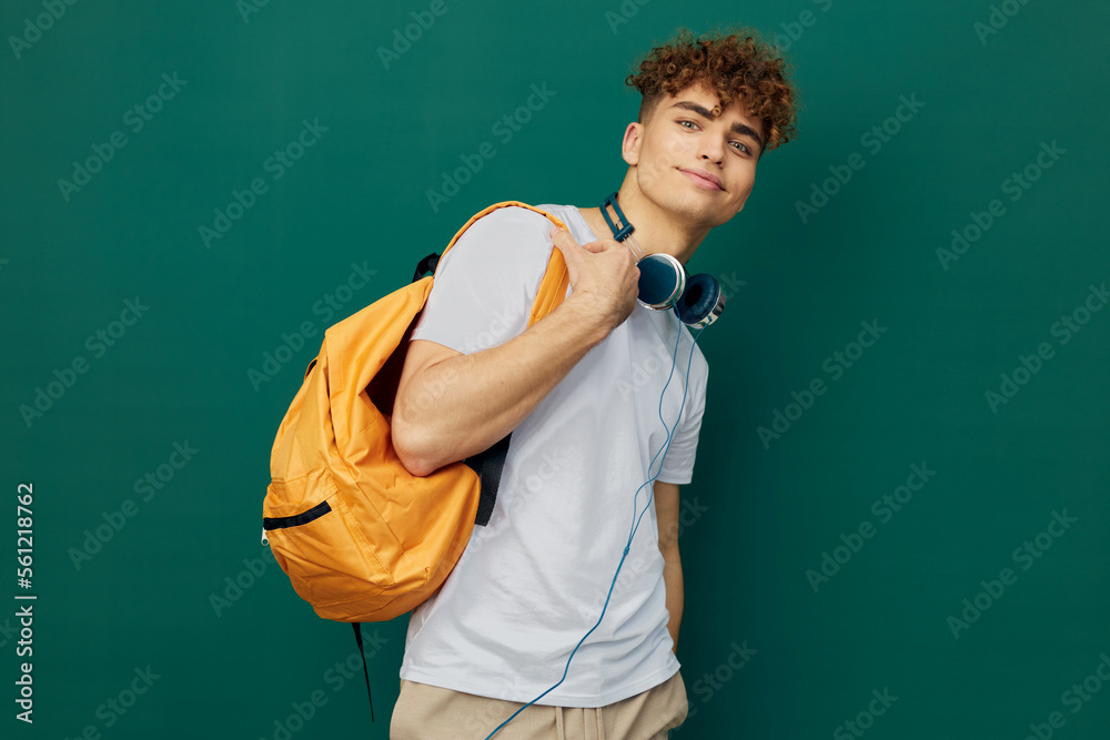 a close horizontal portrait of a handsome man with curly hair standing on a green background in a gray T-shirt, holding headphones around his neck, holding a bright backpack in his hand