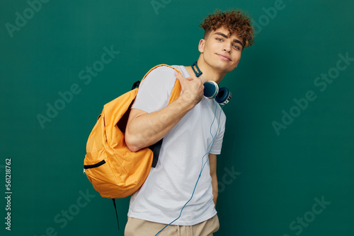 a close horizontal portrait of a handsome man with curly hair standing on a green background in a gray T-shirt, holding headphones around his neck, holding a bright backpack in his hand