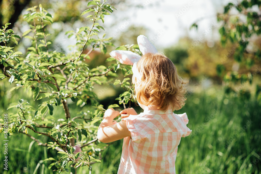 Easter egg hunt. Girl child Wearing Bunny Ears Running To Pick Up Egg In Garden. Easter tradition. Baby with basket full of colorful eggs. Easter egg hunt. Girl child Wearing Bunny Ears Running To Pick Up Egg In Garden. Easter tradition. Baby with basket full of colorful eggs.