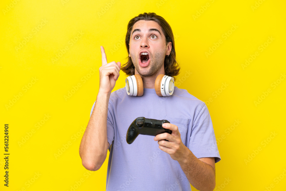 Young handsome caucasian man playing with a video game controller over isolated on yellow background thinking an idea pointing the finger up