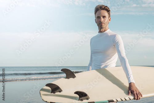 Young handsome surfer guy wearing an empty blank lycra t-shirt and holding a surfboard. Sunlight flare. Surfing mock-up style.