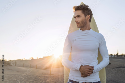 Foto Young handsome surfer guy is standing next to his surfboard and wearing white, empty, blank lycra shirt