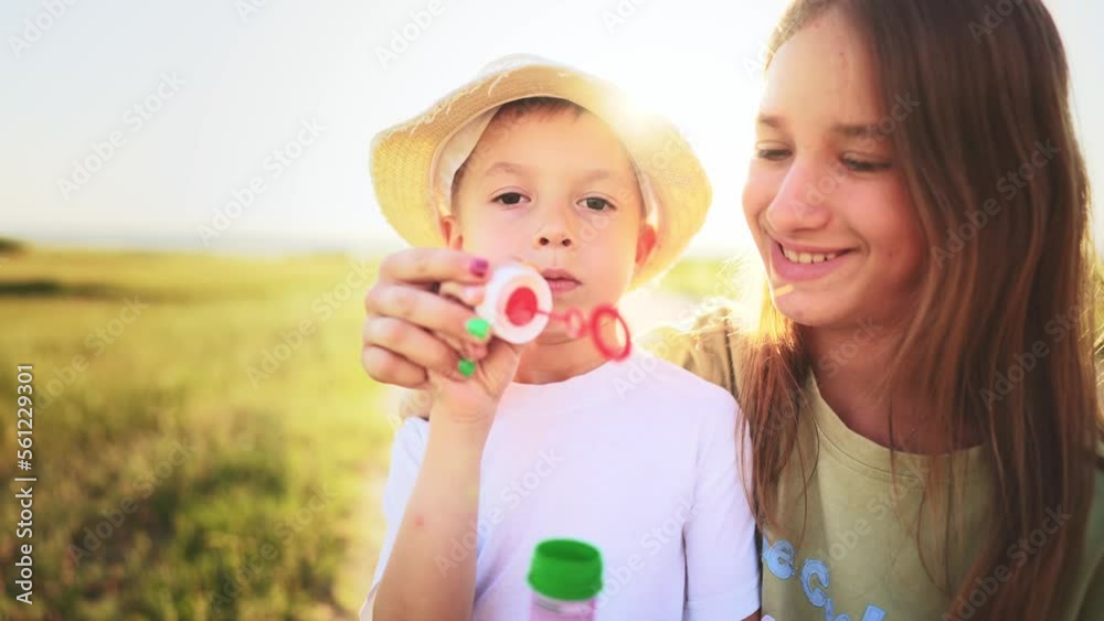 Teenage sister stands and smiles next to little brother who blows and gets soap bubbles in outdoor in field. Family walk on weekend out of town in countryside. Leisure activity. Healthy lifestyle. Teenage sister stands and smiles next to little brother who blows and gets soap bubbles in outdoor in field. Family walk on weekend out of town in countryside. Leisure activity. Healthy lifestyle.