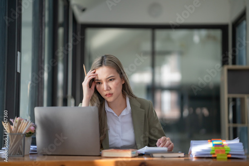 Sad Asian woman looking annoyed and stressed, sitting at the desk, using a laptop, thinking, feeling tired and bored with depression problems