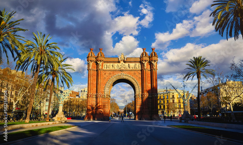Sunrise at Triumphal Arch in Barcelona, Catalonia, Spain. Arc de Triomf boulevard street. Alley with tropical palm trees. Early morning landscape shadows and blue sky clouds. Famous landmark