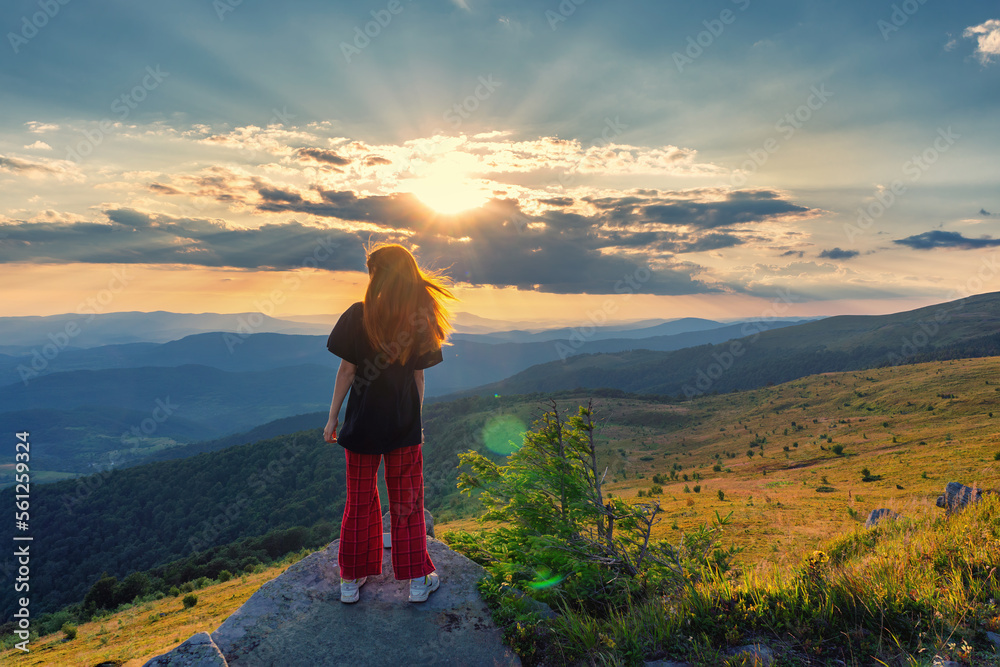 Naklejka premium A young, beautiful girl with flowing red hair against the backdrop of a sunset in the mountains. Beauty stands on a rock