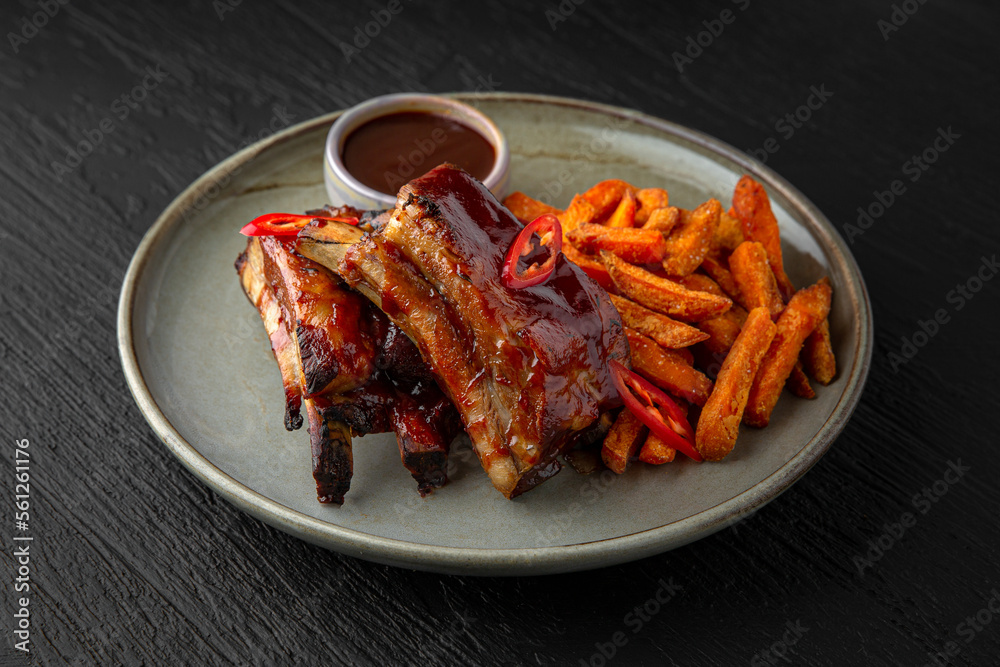 Pork ribs with sweet potato fries in a ceramic plate on a dark textured background. Restaurant menu Isolated on black