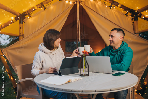 Happy family couple freelancers drink coffee while working laptop on cozy glamping tent in summer evening. Luxury camping tent for outdoor holiday and vacation. Lifestyle concept