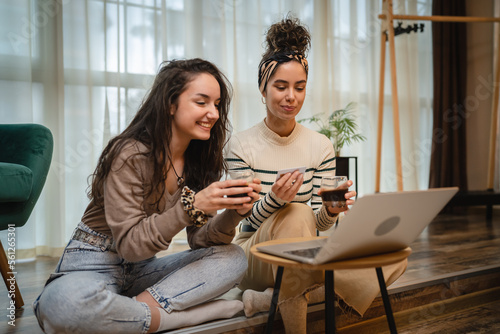 Two friends or sisters or girlfriends are sitting on the ground watching on laptop what to buy online while smiling and talking and drinking coffee in their apartment 