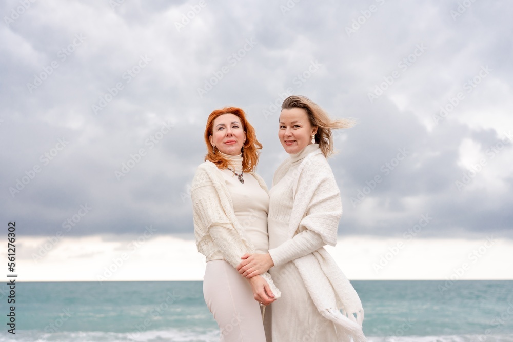 Women sea walk friendship spring. Two girlfriends, redhead and blonde, middle-aged walk along the sandy beach of the sea, dressed in white clothes. Against the backdrop of a cloudy sky and the winter Women sea walk friendship spring. Two girlfriends, redhead and blonde, middle-aged walk along the sandy beach of the sea, dressed in white clothes. Against the backdrop of a cloudy sky and the winter