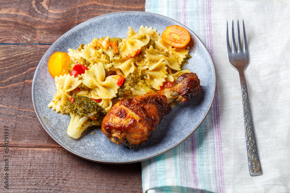 Pasta with vegetables and cherry tomatoes with baked chicken leg on the table next to a light napkin and fork. Pasta with vegetables and cherry tomatoes with baked chicken leg on the table next to a light napkin and fork.