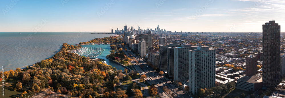 Obraz premium Beautiful cityscape aerial panorama of the Lakeview neighborhood lakefront with autumn foliage surrounding Belmont Harbor and the Chicago skyline in the distance on a sunny day.