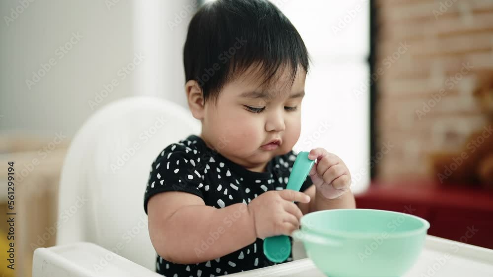 Adorable hispanic baby sitting on highchair holding spoon at home Adorable hispanic baby sitting on highchair holding spoon at home