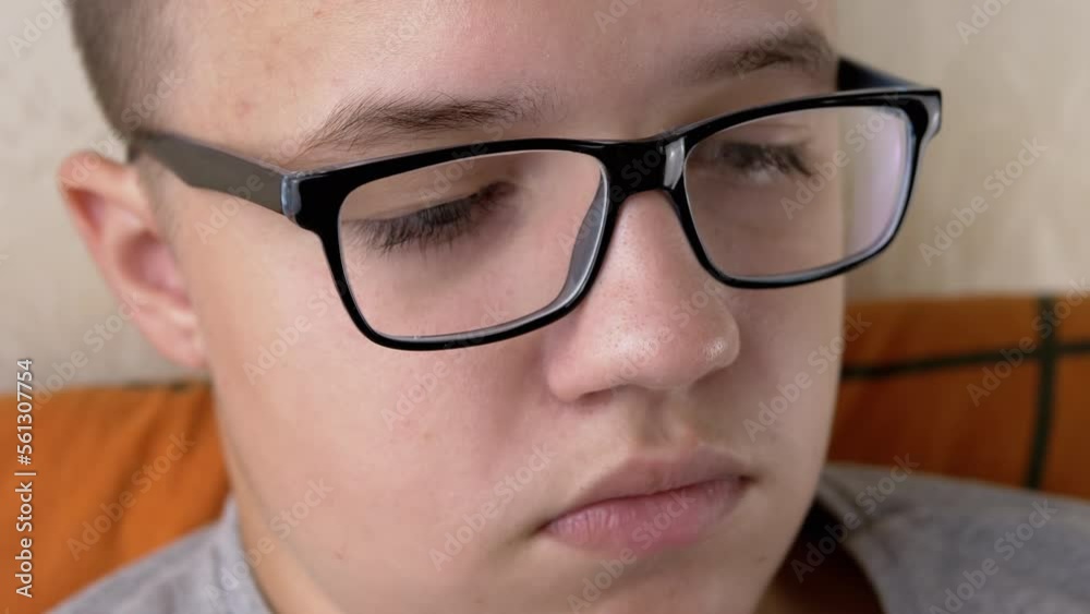 Close-up Face of an Offended Serious Schoolboy with Glasses, with Full Lips. Thinking tired teenager with sad downcast eyes, thick long eyelashes in a room. Portrait, body part. Child, boy. Emotions.
