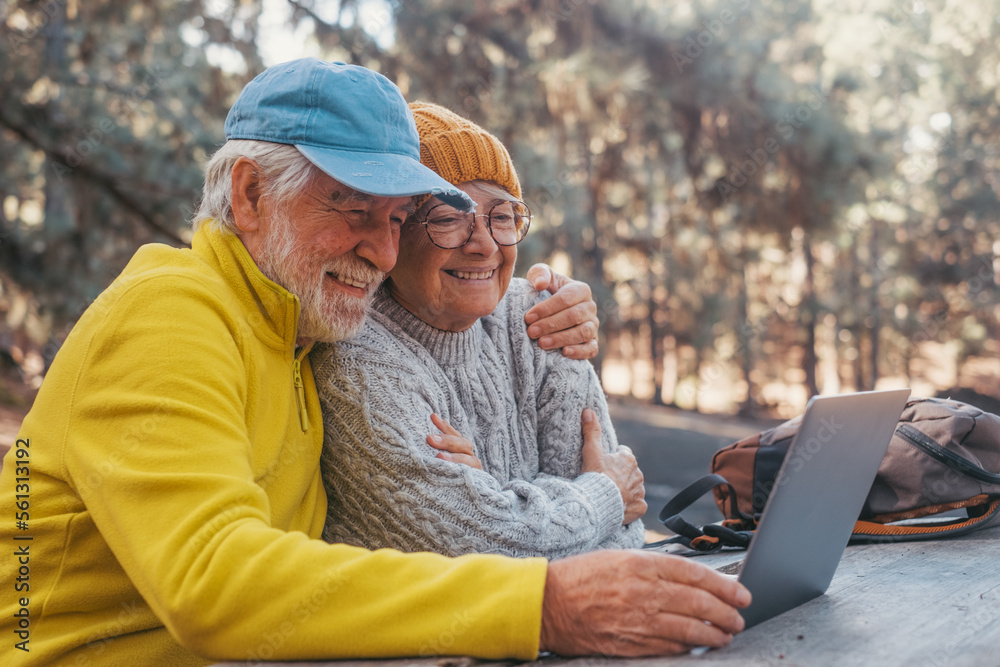 Φωτογραφία Head shot portrait close up of cute couple of old middle age people using computer pc outdoors sitting at a wooden table in the forest of mountain in nature with trees around them