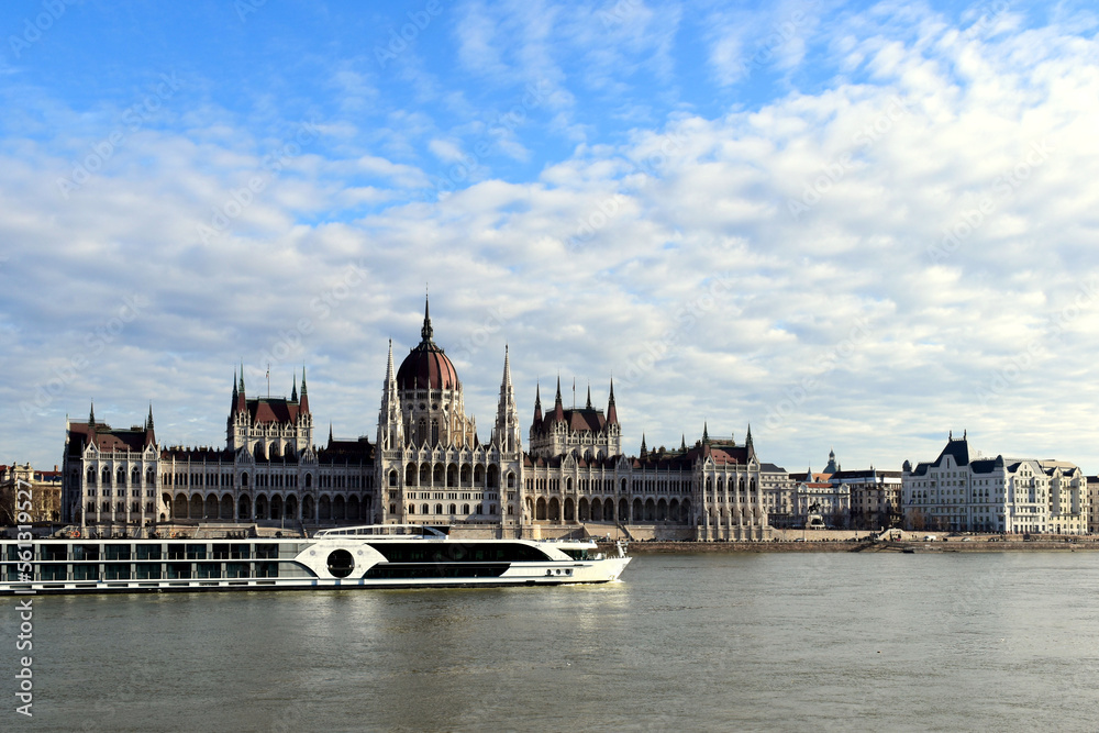 the Hungarian Parliament in Neo-Gothic style from across the Danube in panoramic view. white tour and hotel boat going by. blue sky. Famous and popular Budapest landmark and attraction. City skyline.