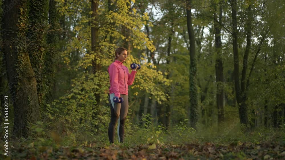 Slow-motion shot of a beautiful young woman with earphones exercising with dumbbells in the forest at sunset. Healthy lifestyle concept Slow-motion shot of a beautiful young woman with earphones exercising with dumbbells in the forest at sunset. Healthy lifestyle concept