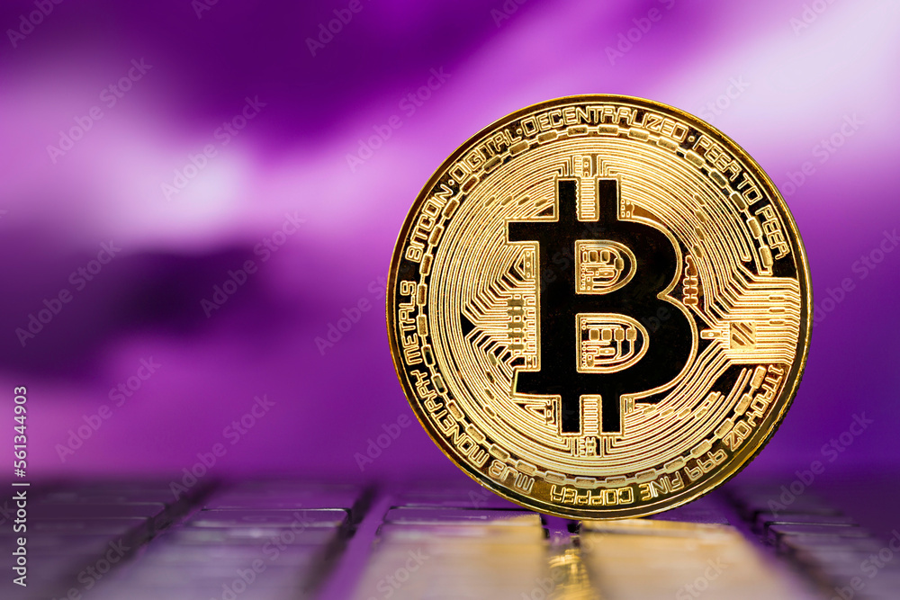 Golden metallic Bitcoin coin standing on a laptop keyboard against the background of a blurred  multicolored sky 