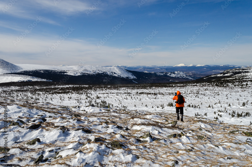 Obraz premium Mountain winter landscape. A tourist with a backpack walks through a snow-covered field. In the background, snowy mountains, blue sky.