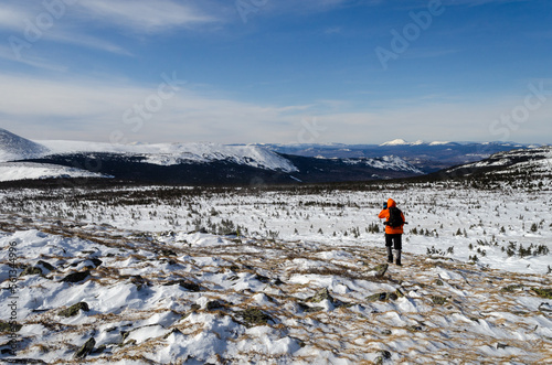 Mountain winter landscape. A tourist with a backpack walks through a snow-covered field. In the background, snowy mountains, blue sky.