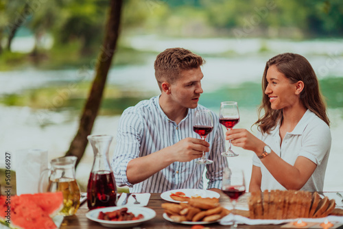 Happy couple toasting red wine glass while having picnic french dinner party outdoor during summer holiday vacation near the river at beautiful nature