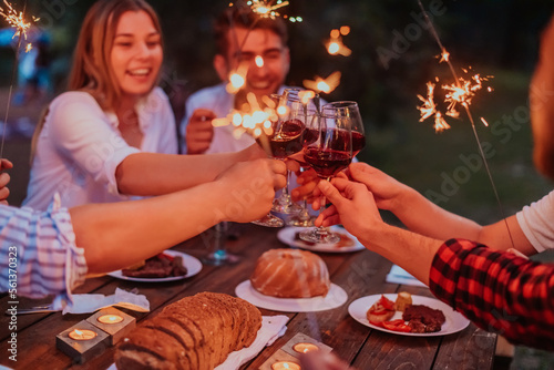 Group of happy friends celebrating holiday vacation using sprinklers and drinking red wine while having picnic french dinner party outdoor near the river on beautiful summer evening in nature