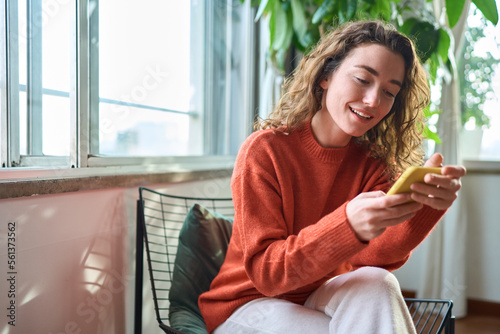 Fotografie, Tablou Smiling young woman sitting on chair holding mobile phone using cellphone device, looking at smartphone, checking modern apps, texting messages, browsing internet doing shopping relaxing at home