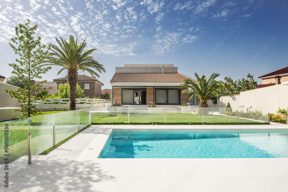 Beautiful pool in a farm with marble floors, glass railing, some palm trees in the garden and a house in the background