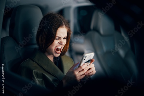 a close horizontal portrait of a stylish, luxurious woman in a leather coat sitting in a black car at night in the passenger seat, emotionally looking into her smartphone with her mouth wide open © SHOTPRIME STUDIO