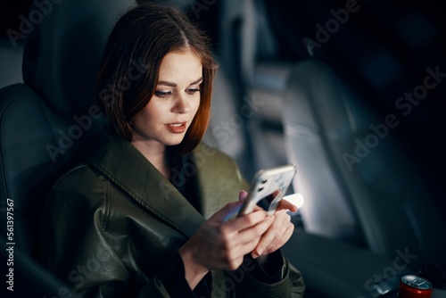 a close horizontal portrait of a stylish, luxurious woman in a leather coat sitting in a black car at night in the passenger seat, looking at her phone during the trip