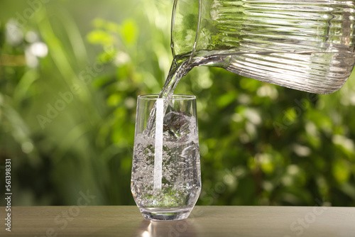 Pouring water from jug into glass on wooden table outdoors, closeup