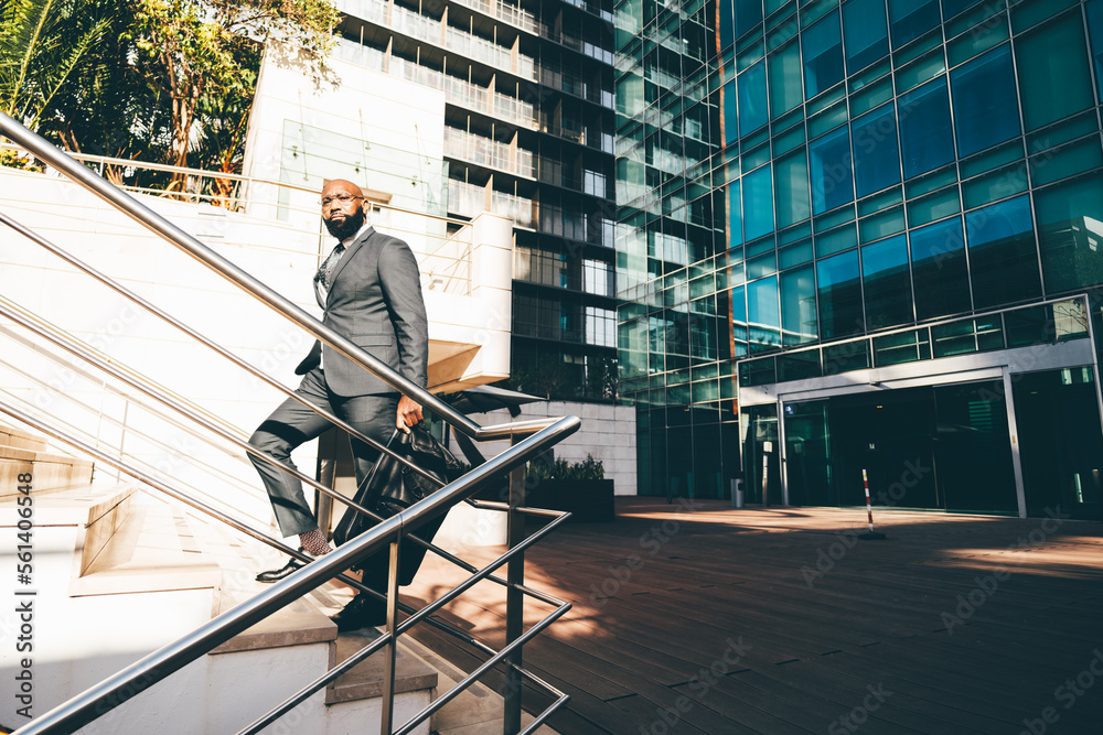 A black white-collar employee moving up the staircase, a metallic railing next to him, the office building with blue windows behind him and he is carrying a weekender bag, wearing dark grey suit