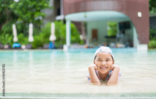 Asian child smile or kid girl wear swimsuit cap to happy lying down relax on swimming pool in water park or person learning swim to sports exercise on summer school or holiday vacation travel at hotel